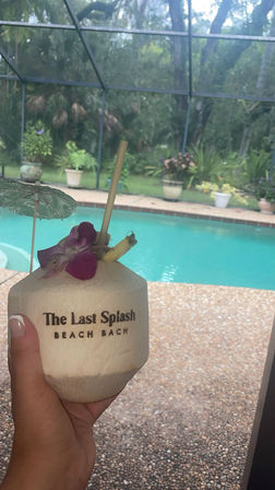 Hand holding a tropical coconut drink with orchid garnish, straw and mini umbrella in front of a screened backyard pool and potted plants