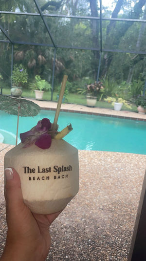 Hand holding a tropical coconut drink with orchid garnish, straw and mini umbrella in front of a screened backyard pool and potted plants