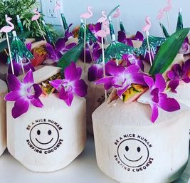 Fresh young coconuts turned into tropical drinks, topped with bright purple orchid flowers, green paper tiki umbrellas, pink flamingo picks and a stamped smiley-face logo for a festive, beachy presentation.