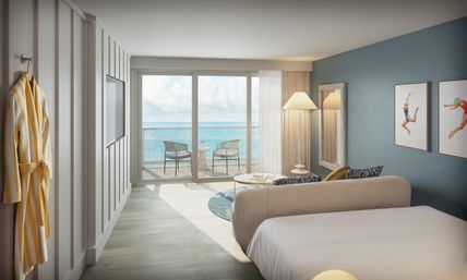 Oceanfront beachside hotel room with king bed and sofa, sliding glass doors to a balcony with two chairs and a table overlooking turquoise sea, striped yellow robe on a hook, blue accent wall and coastal artwork.