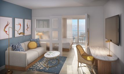 Bright ocean-view hotel suite with balcony, modern coastal decor: cream sofa against blue wall with diving art, marble coffee table, gold lamps, rattan desk and wall TV, partitioned bedroom.