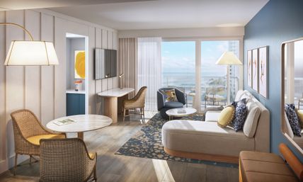 Sunlit coastal hotel suite living room with ocean-view balcony, blue accent wall, chaise lounge with patterned pillows, wicker dining chairs, round marble tables, small work desk and wall-mounted TV.