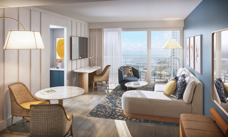 Sunlit coastal hotel suite living room with ocean-view balcony, blue accent wall, chaise lounge with patterned pillows, wicker dining chairs, round marble tables, small work desk and wall-mounted TV.