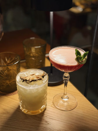 Two craft cocktails on a wooden table under warm lamp light — a pink frothy coupe garnished with mint and a pale crushed-ice drink in a patterned rocks glass topped with dried fruit.