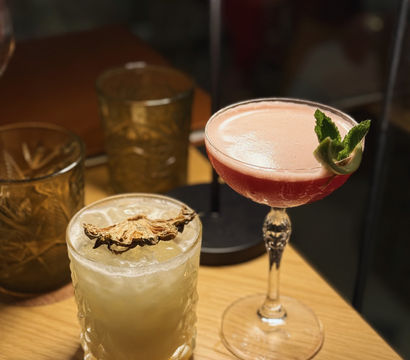 Two craft cocktails on a wooden table under warm lamp light — a pink frothy coupe garnished with mint and a pale crushed-ice drink in a patterned rocks glass topped with dried fruit.