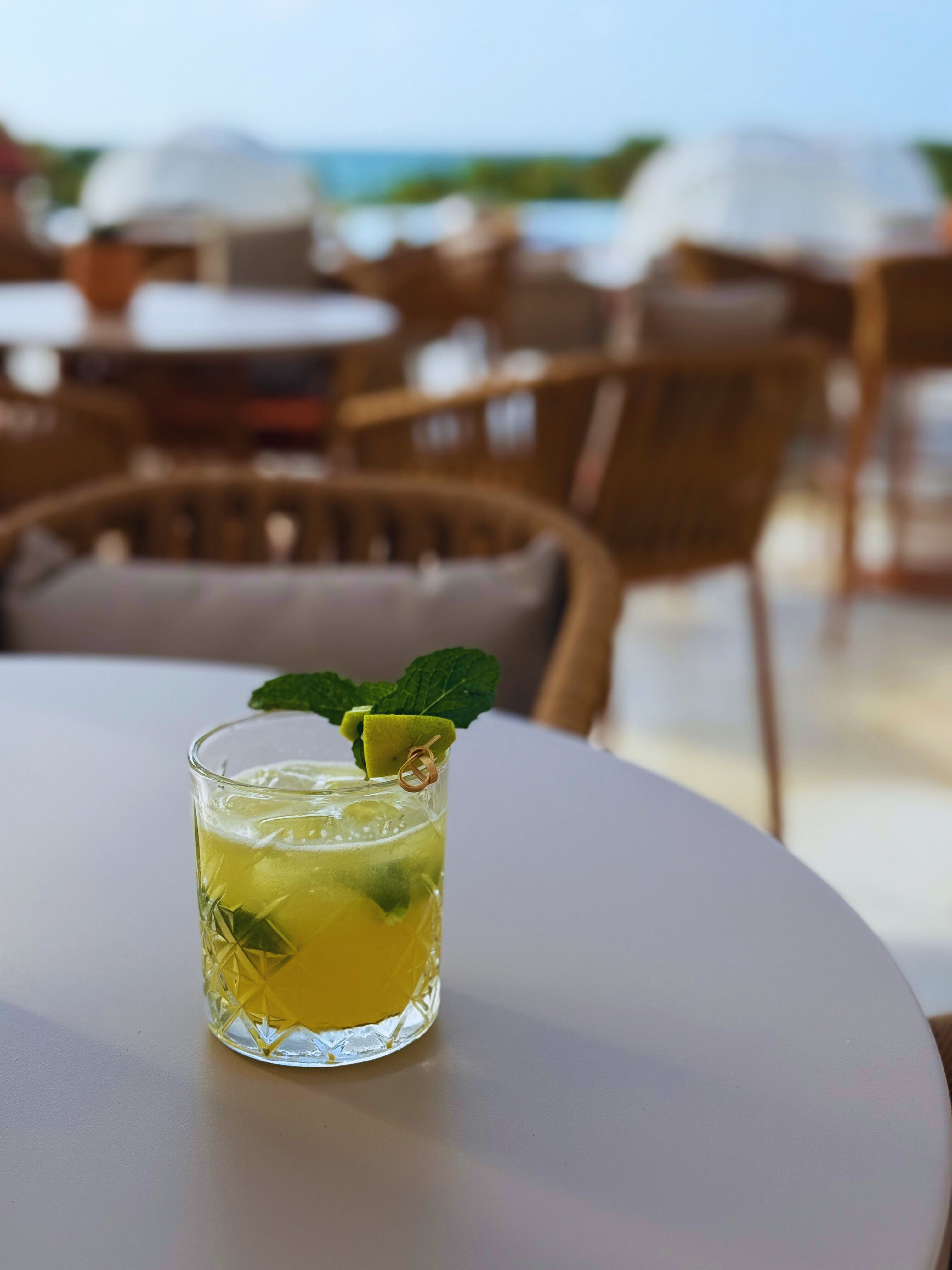 Refreshing lime-and-mint cocktail in a cut-glass tumbler on a white table at a sunny outdoor patio with wicker chairs and a blurred sea view