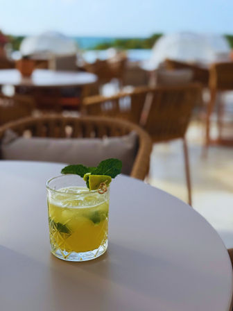 Refreshing lime-and-mint cocktail in a cut-glass tumbler on a white table at a sunny outdoor patio with wicker chairs and a blurred sea view