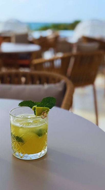 Refreshing lime-and-mint cocktail in a cut-glass tumbler on a white table at a sunny outdoor patio with wicker chairs and a blurred sea view