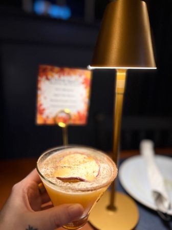 Hand holding a frothy peach cocktail in a rimmed coupe glass topped with dried fruit, on a dim restaurant table beside a brass table lamp and fall-themed menu.