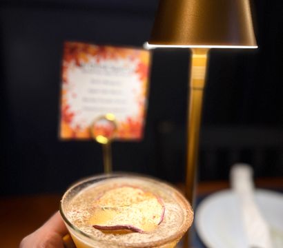 Hand holding a frothy peach cocktail in a rimmed coupe glass topped with dried fruit, on a dim restaurant table beside a brass table lamp and fall-themed menu.