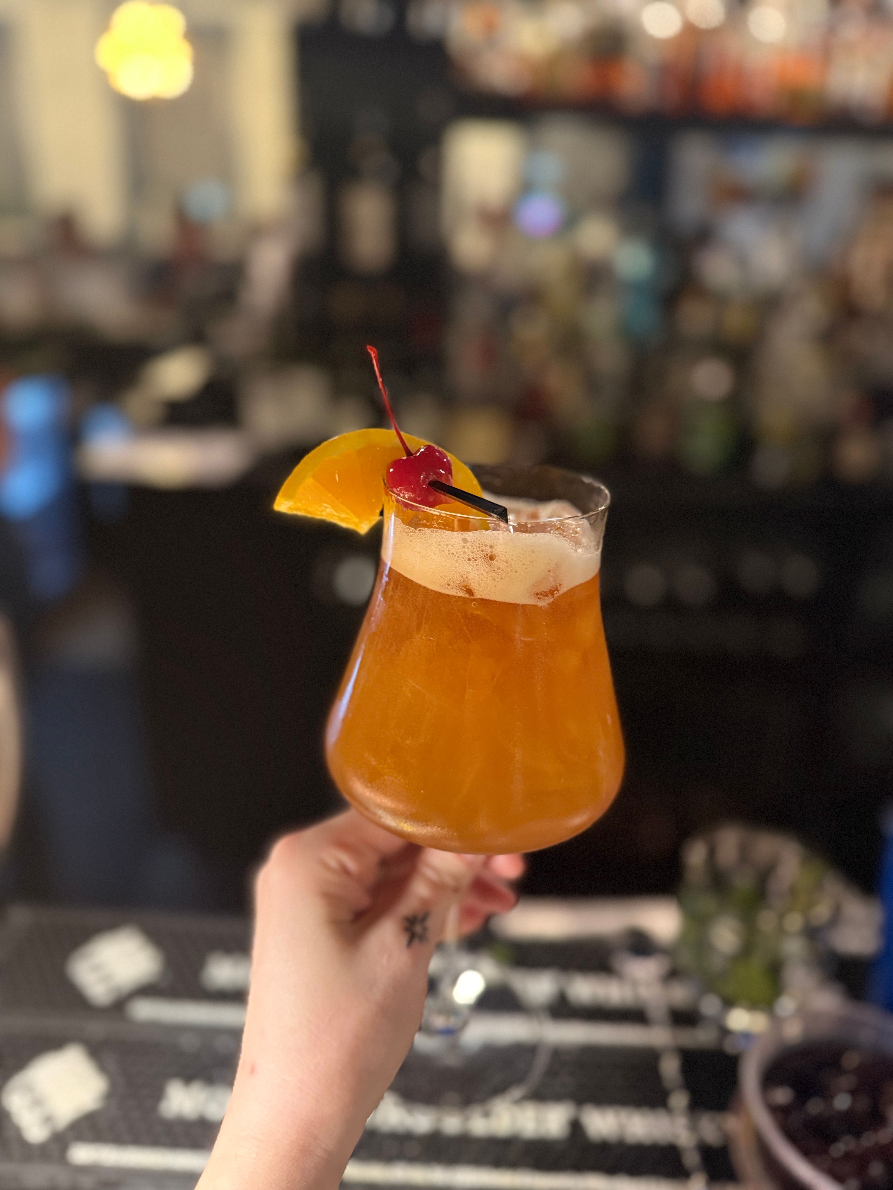 Hand holding amber craft cocktail in a tulip glass with frothy top, garnished with an orange slice and maraschino cherry at a dimly lit bar