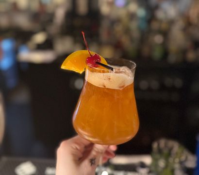 Hand holding amber craft cocktail in a tulip glass with frothy top, garnished with an orange slice and maraschino cherry at a dimly lit bar