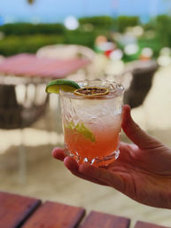Hand holding pink seaside cocktail in a cut-glass tumbler with crushed ice, lime wedge and dried citrus wheel, blurred tropical beach tables and ocean view in background.