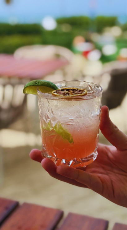 Hand holding pink seaside cocktail in a cut-glass tumbler with crushed ice, lime wedge and dried citrus wheel, blurred tropical beach tables and ocean view in background.