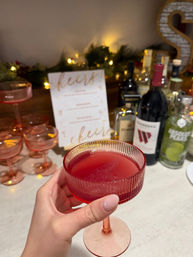 Hand holding a pink coupe glass filled with a deep red cocktail on a home bar counter, pink glassware stacked beside bottles, twinkling lights and a “cheers” menu in the background — cozy holiday cocktail setup.