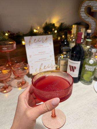 Hand holding a pink coupe glass filled with a deep red cocktail on a home bar counter, pink glassware stacked beside bottles, twinkling lights and a “cheers” menu in the background — cozy holiday cocktail setup.