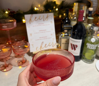 Hand holding a pink coupe glass filled with a deep red cocktail on a home bar counter, pink glassware stacked beside bottles, twinkling lights and a “cheers” menu in the background — cozy holiday cocktail setup.