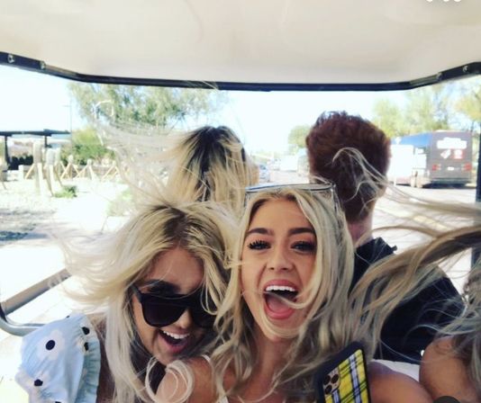 Two friends laughing with wind-blown hair riding in an open-air golf cart on a sunny desert road, one holding a smartphone