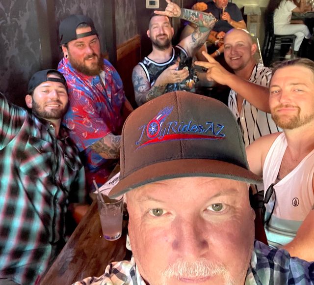 Group selfie of six men at a lively bar, close-up foreground of a man in a baseball cap, friends smiling and posing, visible tattoos and casual summer shirts, drinks on a wooden table — energetic pub nightlife scene