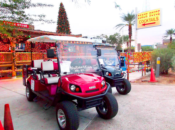 Festive red and black golf carts decked in string lights parked outside a holiday-lit outdoor bar with a tall Christmas tree, palm trees and colorful decorations at dusk.