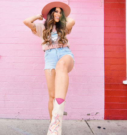 Smiling woman in a wide-brim hat, graphic tee and denim shorts playfully kicking toward camera in embroidered cowboy boots against a bright pink brick wall mural — upbeat street-style portrait.