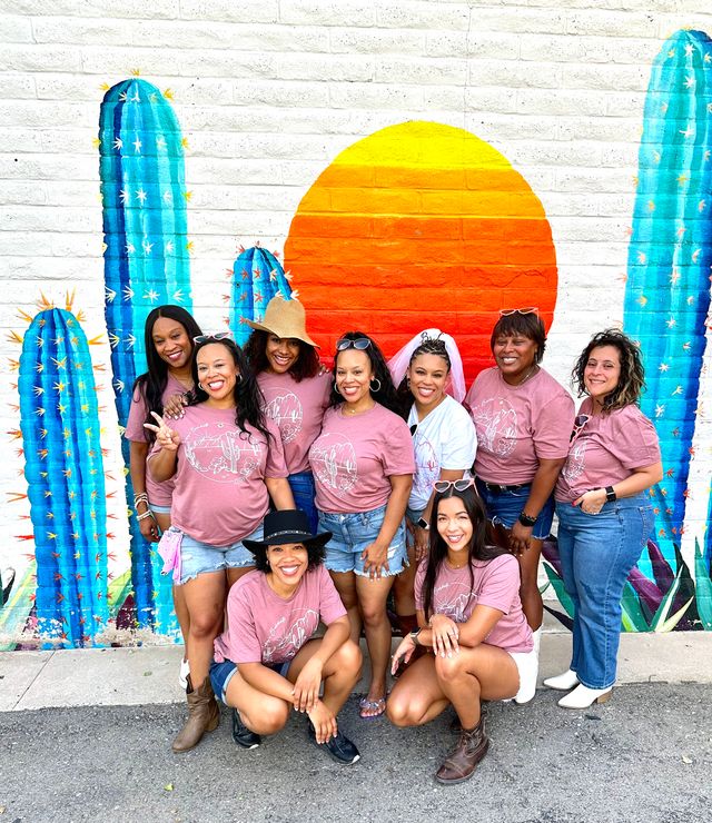 Group of women in matching pink tees and denim posing for a bachelorette-style photo in front of a vibrant cactus and orange sunset mural on a white brick wall