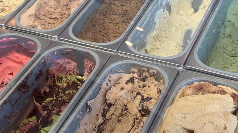 Glass display case of gelato and ice cream tubs in a shop, showing colorful flavors—pink, mint green, chocolate brown and creamy beige swirled with chocolate ribbons and chunks.
