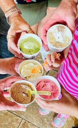 Overhead shot of five hands holding paper cups of colorful ice cream—pistachio green, chocolate, pink strawberry and two creamy vanilla varieties with bright plastic spoons—family enjoying treats at an ice cream shop.