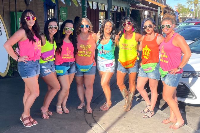Group of women on a sunny downtown street wearing neon tank tops, denim shorts, mirrored sunglasses and colorful fanny packs, posing for a fun bachelorette party photo outside shops.