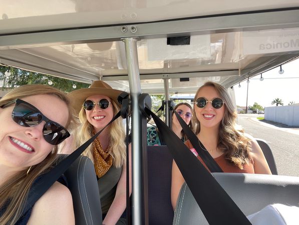 Four smiling friends wearing sunglasses and seatbelts riding in an open golf cart on a sunny street with palm trees.