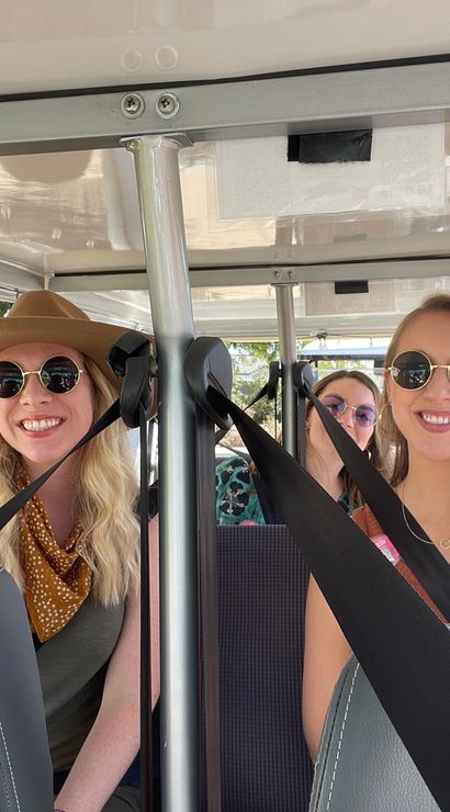 Four smiling friends wearing sunglasses and seatbelts riding in an open golf cart on a sunny street with palm trees.