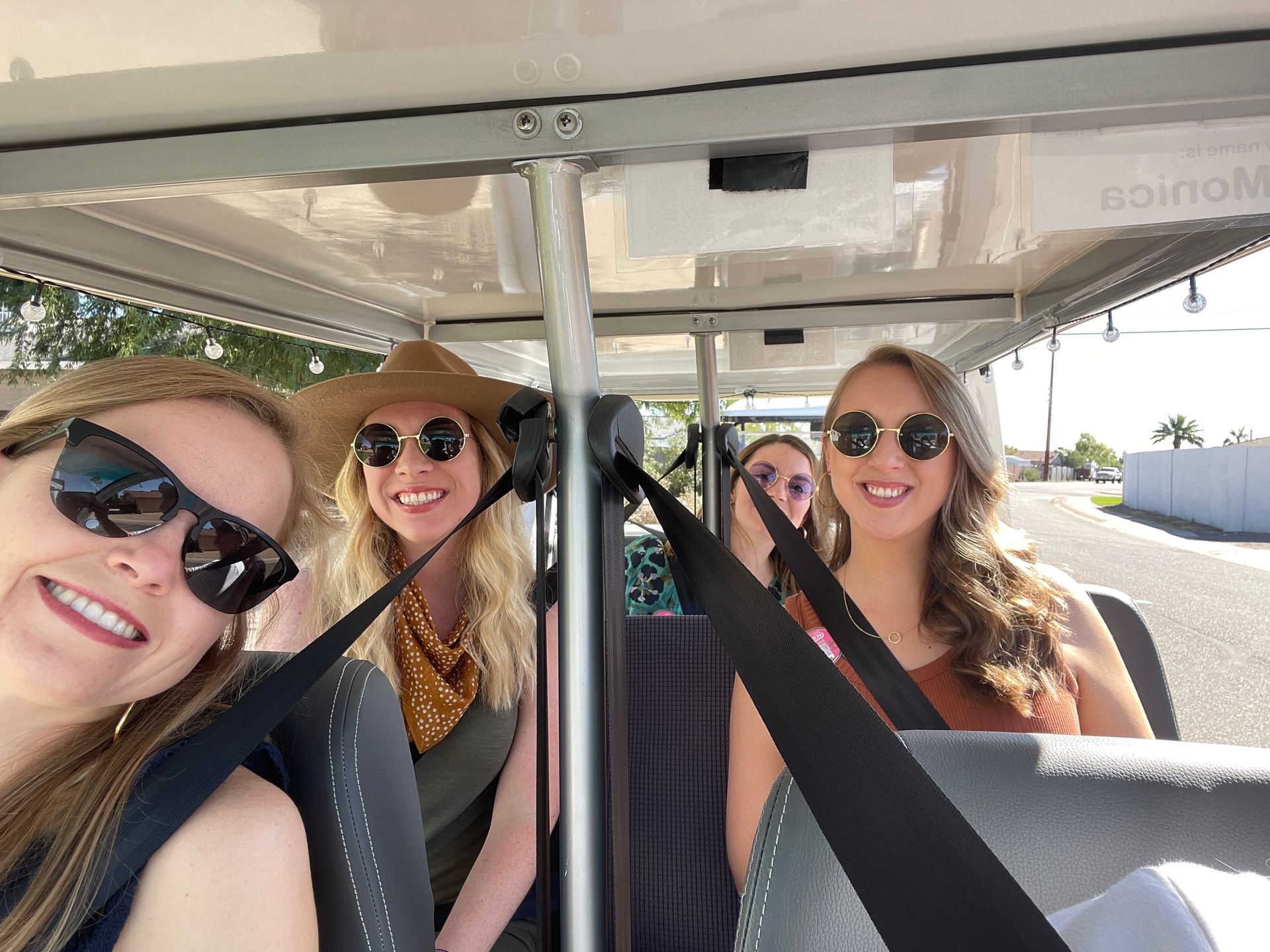 Four smiling friends wearing sunglasses and seatbelts riding in an open golf cart on a sunny street with palm trees.