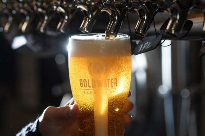 Hand holding a pint glass under a beer tap as golden draft beer pours in, forming a frothy head with a warm backlit glow at a bar.
