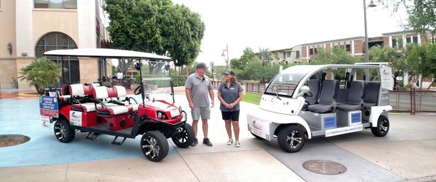 Two electric shuttle carts, one red and one white, parked on a sunny downtown plaza with two drivers chatting between them and mixed-use buildings and trees in the background.