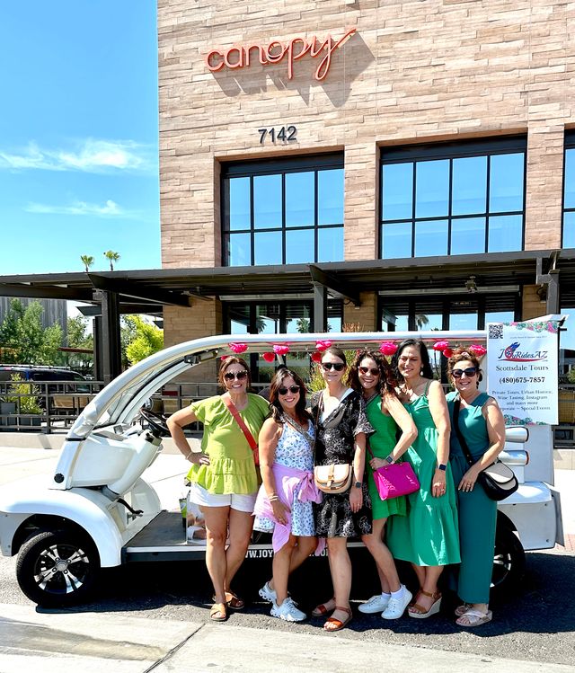 Six women smiling and posing by a white electric tour cart in front of a modern stone building on a sunny downtown Scottsdale, Arizona street — daytime tour group.