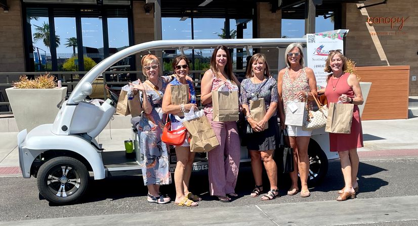 Six women smiling with paper shopping bags beside a white electric shuttle cart at a sunny outdoor shopping center in Scottsdale, Arizona, with palm trees in the background.