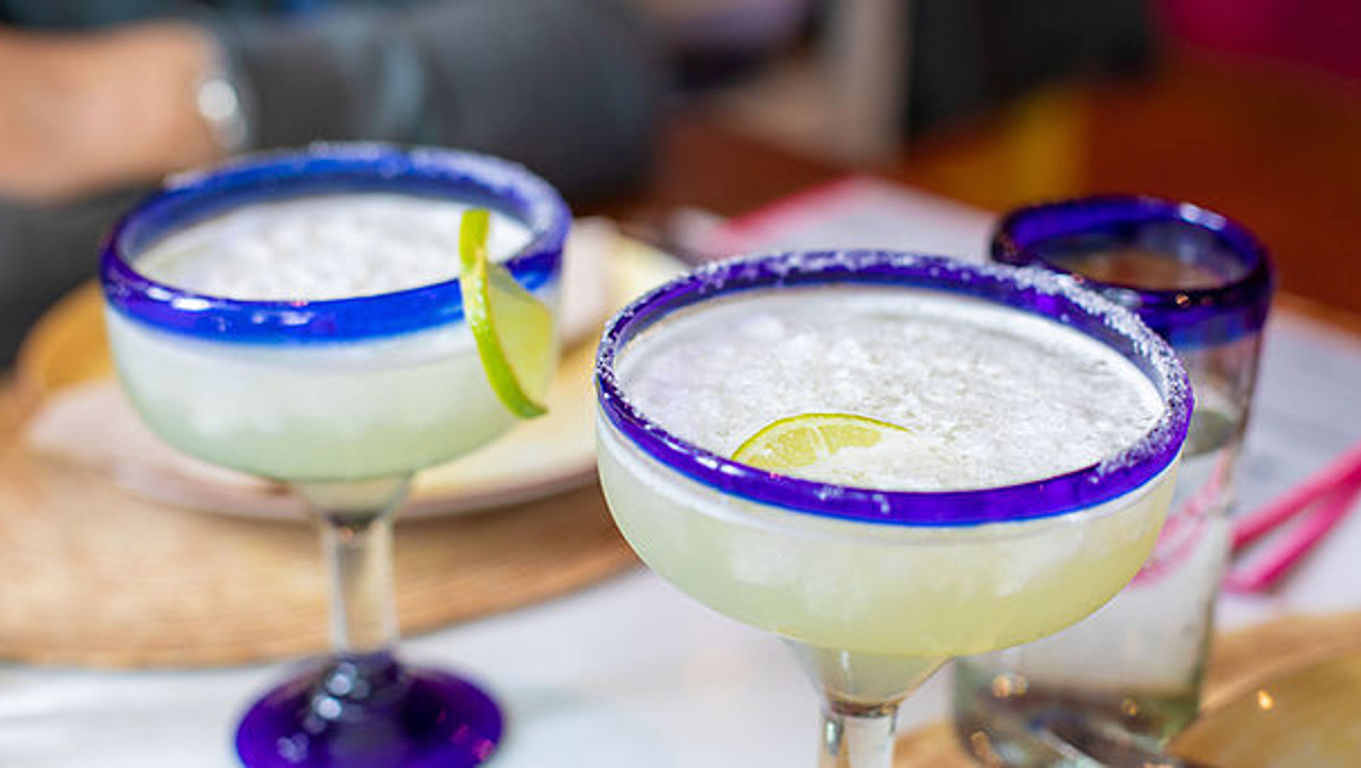 Two chilled lime margaritas with crushed ice and salted rims served in blue-rimmed glasses, garnished with lime wedges on a restaurant table.