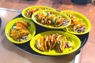 Overhead shot of vibrant green plates holding rows of crispy Mexican tacos filled with shredded lettuce, purple cabbage, crema and salsa, arranged on a black serving tray.
