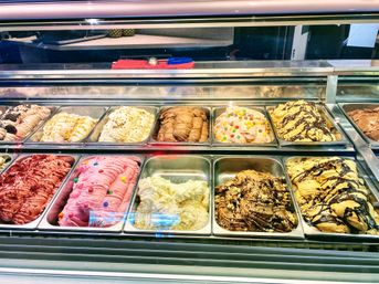 Glass-front gelato display case with colorful metal pans of creamy ice cream—pink and red fruity tubs, vanilla and chocolate swirls, candy-topped and chocolate-drizzled varieties in a dessert shop counter.