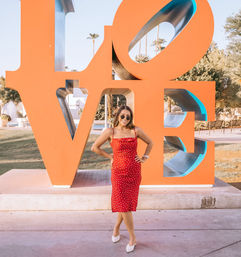 Woman in a red polka-dot dress and sunglasses posing in front of a giant orange LOVE sculpture with palm trees and a fountain in a sunny Palm Springs park