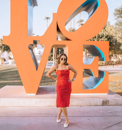 Woman in a red polka-dot dress and sunglasses posing in front of a giant orange LOVE sculpture with palm trees and a fountain in a sunny Palm Springs park