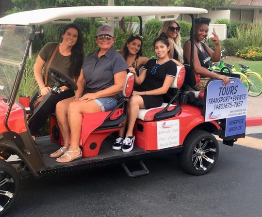 Six smiling women enjoying a red tour-style golf cart parked on a sunny suburban street, with a visible 'Tours' sign and rental bikes in the background.