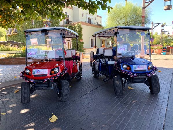 Two festive red and blue golf carts decorated with patriotic tinsel parked under a shade tree on a sunny Southwestern plaza with stucco buildings and desert landscaping