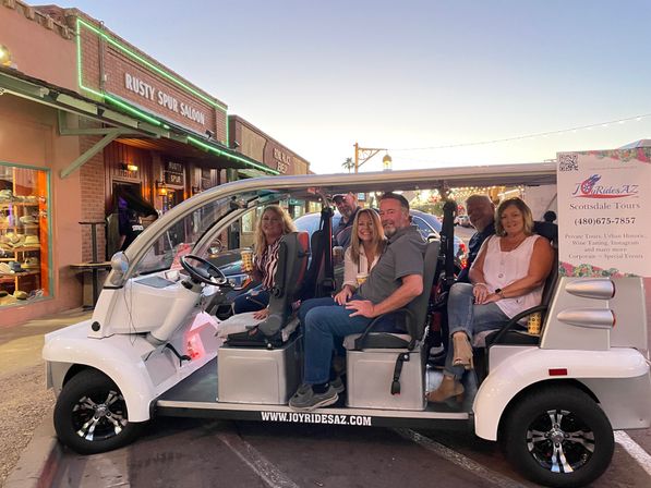 Cheerful adults seated in a white open-air electric tour shuttle on a lively western-style downtown street at dusk in Scottsdale, Arizona, with neon saloon signage and string lights in the background.
