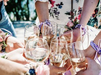 Friends toasting outdoors with white wine in stemmed glasses, wearing floral dresses and pink corsages tied with purple ribbons.