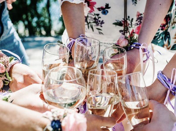 Friends toasting outdoors with white wine in stemmed glasses, wearing floral dresses and pink corsages tied with purple ribbons.