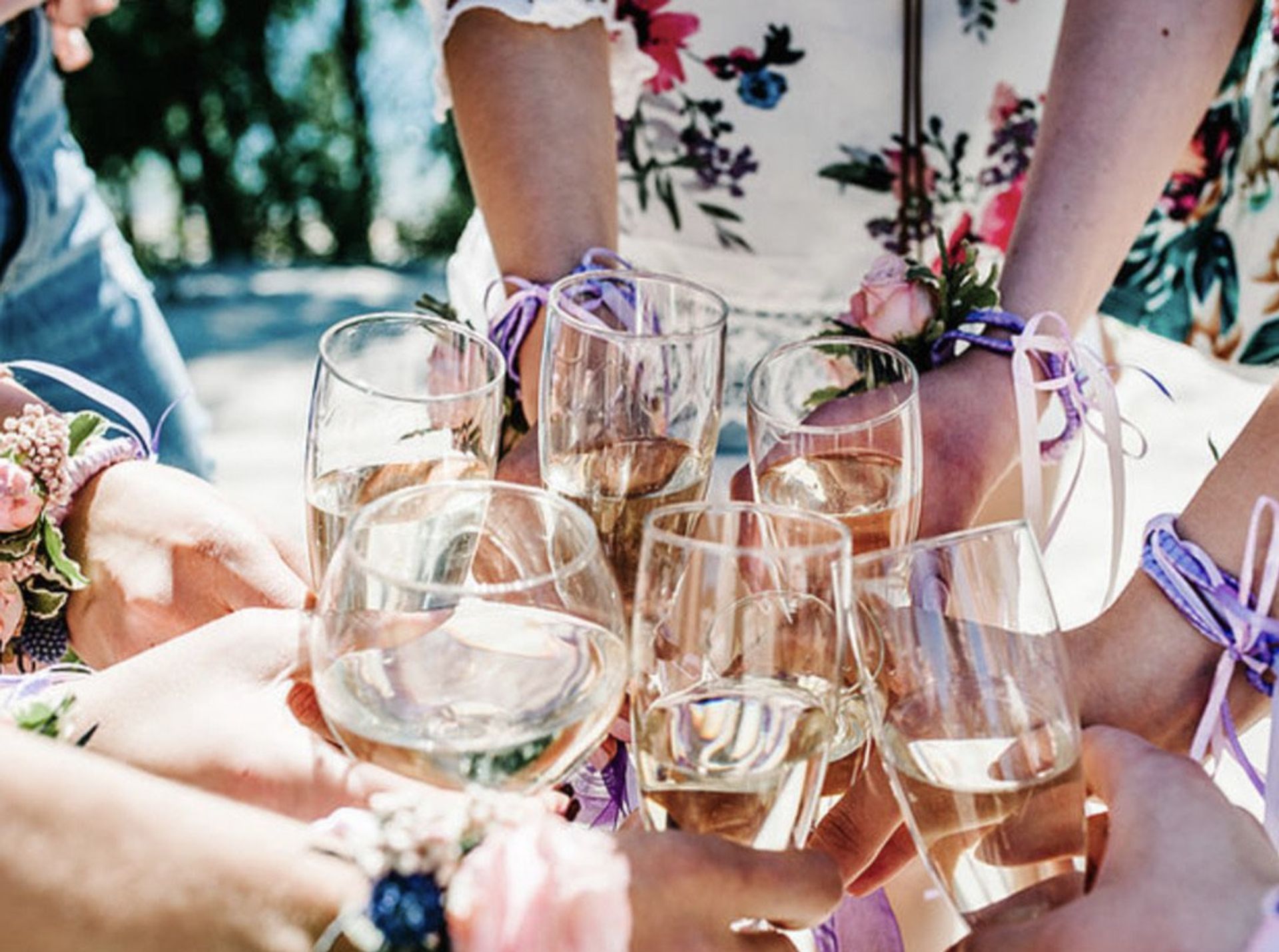 Friends toasting outdoors with white wine in stemmed glasses, wearing floral dresses and pink corsages tied with purple ribbons.