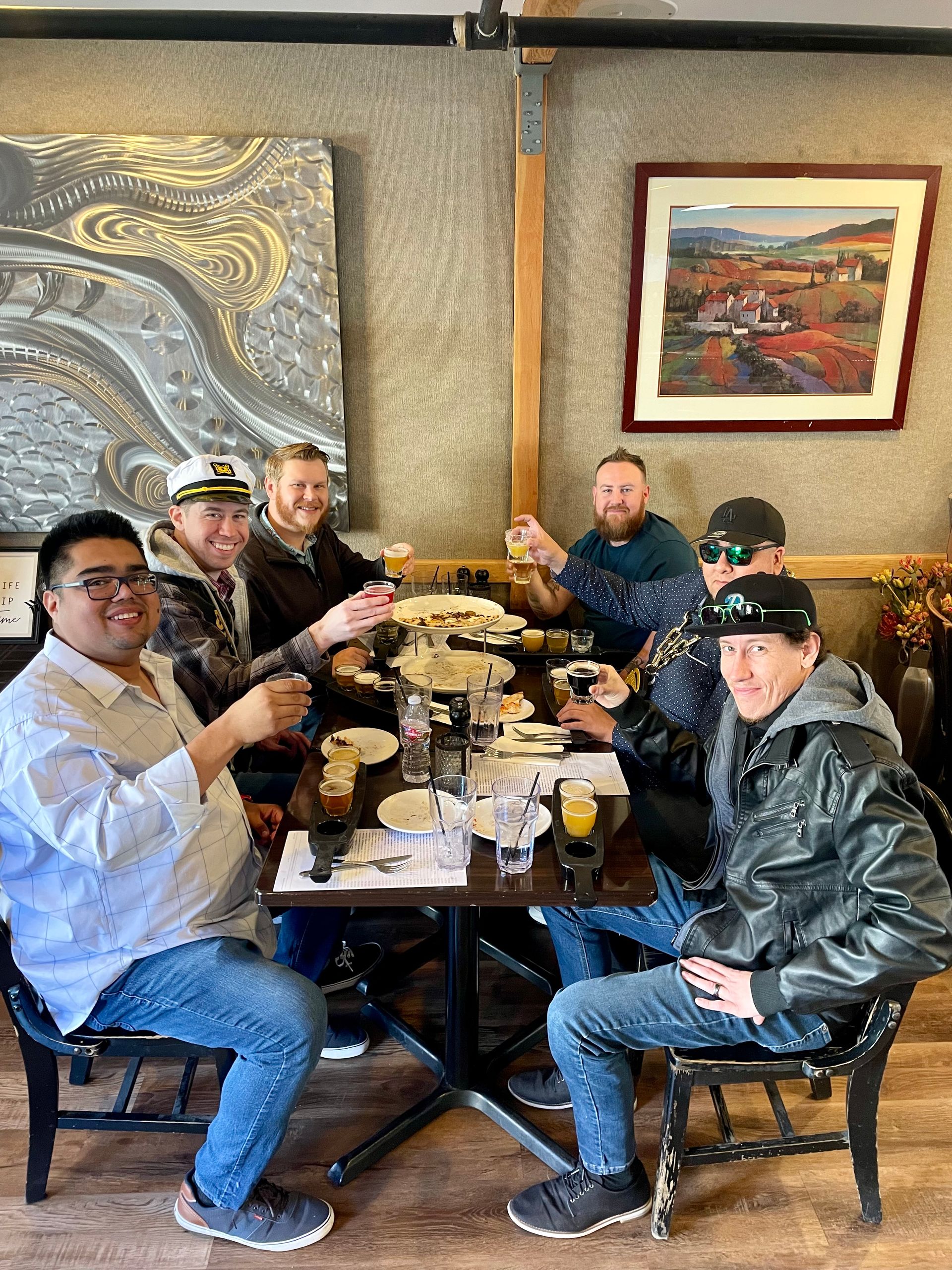 Six friends toasting with small beers over craft-beer flights at a cozy indoor restaurant table with plates and wall art.