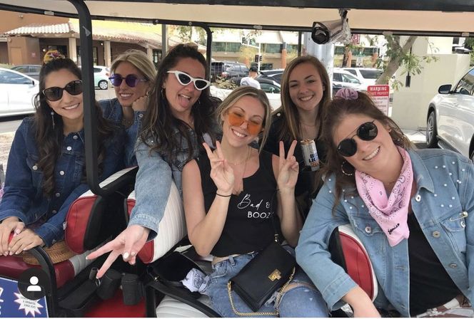 Six friends in a golf cart on a sunny street, wearing sunglasses and denim, smiling and flashing peace signs, one holding a canned drink and another wearing a pink bandana.