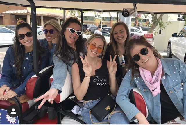 Six friends in a golf cart on a sunny street, wearing sunglasses and denim, smiling and flashing peace signs, one holding a canned drink and another wearing a pink bandana.